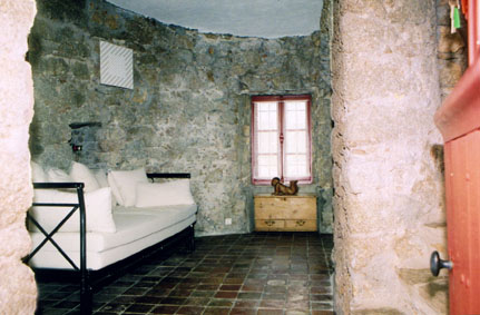 Ground floor of Moulin Tower viewed through the doorway. There is a wrought iron sofa bed with white mattress and cushions. The floor is tiled and the walls are natural stone. There is a small window opposite the door.